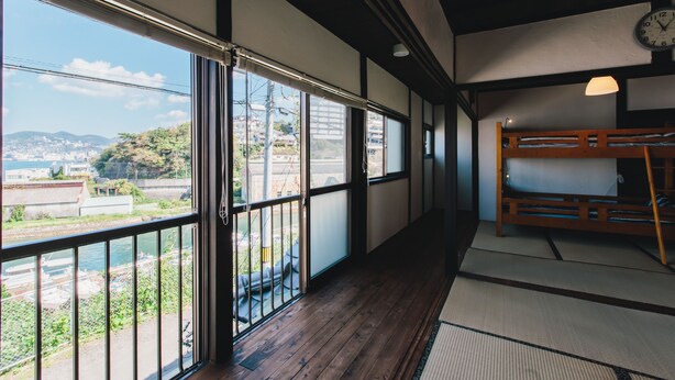 A room with a wooden floor, tatami mat flooring, a bunk bed, and large windows offering a view of a cityscape and water. - Hostel Dockhouse (Nagasaki)