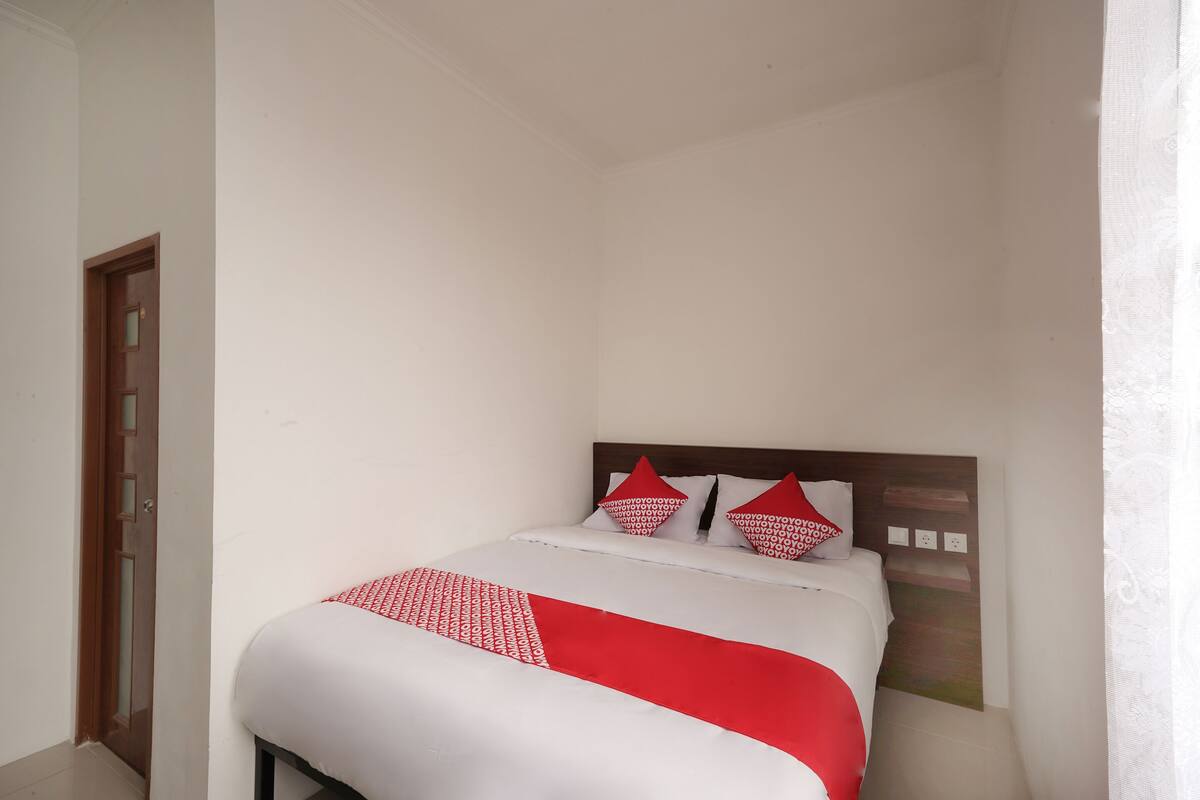 A small, neatly made bed with a white and red bedspread and matching pillows, a wooden headboard, and a plain white wall.