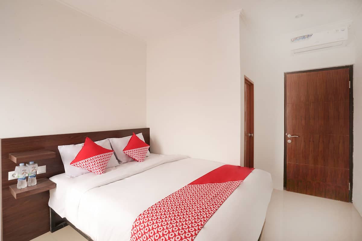 A hotel room with a bed, pillows, a nightstand, a water bottle, a red throw, a wooden door, and an air conditioning unit.