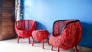 Dos sillas rojas de mimbre con detalles negros y un pequeño reposapiés, ubicados frente a una pared azul y una puerta de madera.