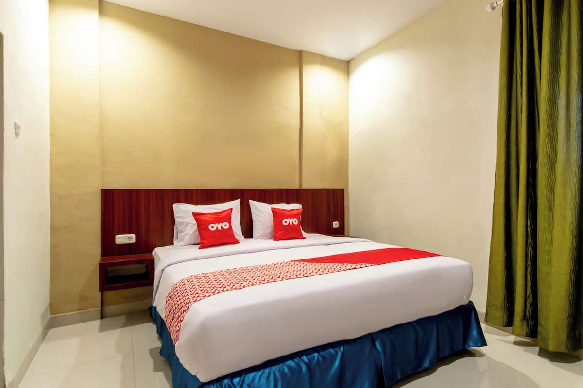 A hotel room with a large bed, two pillows with 'OYO' branding, a red and white bedspread, a wooden headboard, a nightstand, and a green curtain.