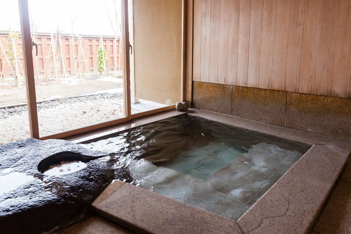 A hot spring bath with a stone border, wooden walls, and a sliding glass door leading outside.