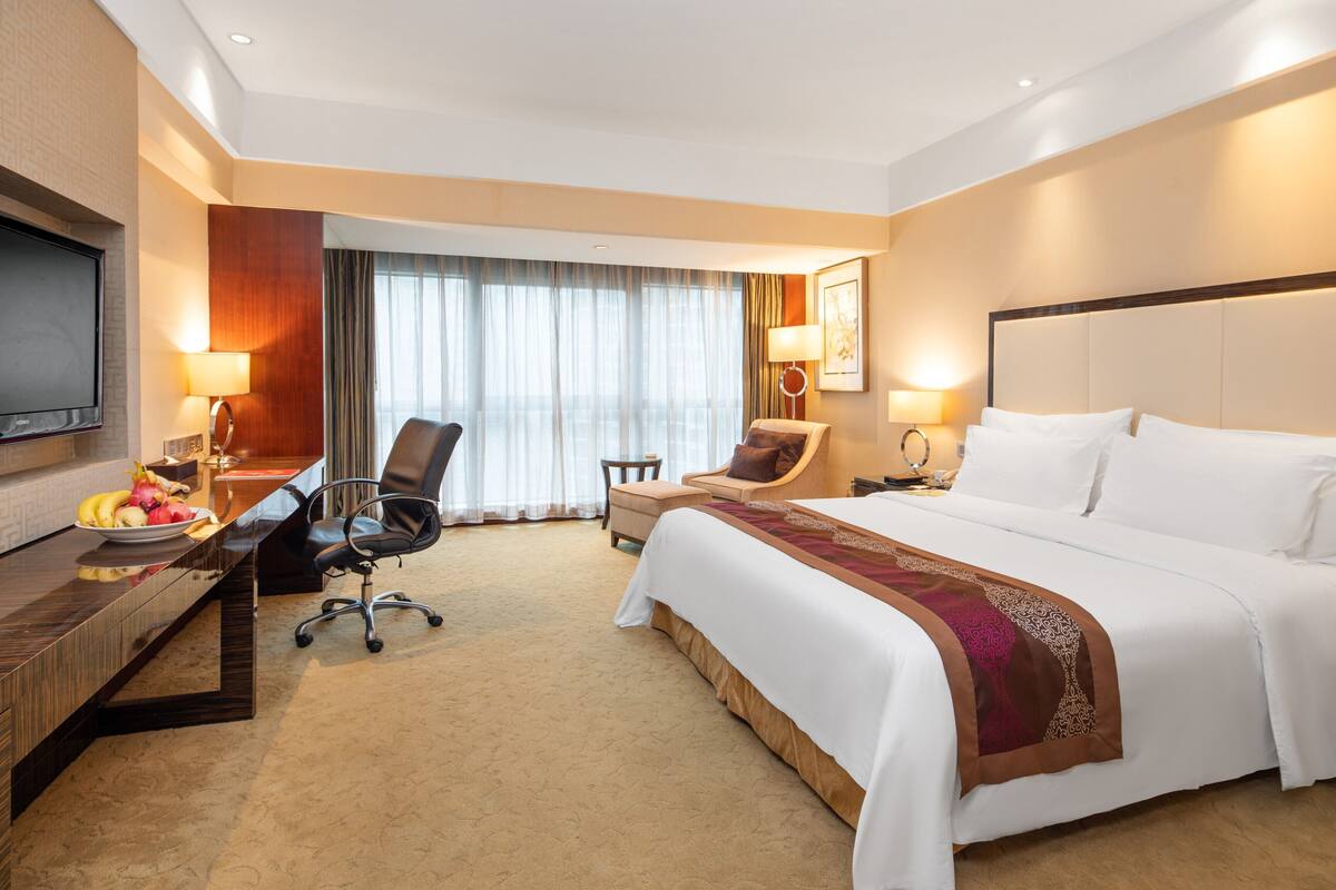 A hotel room with a large bed, a desk with a computer, a chair, a television, and a window with curtains.