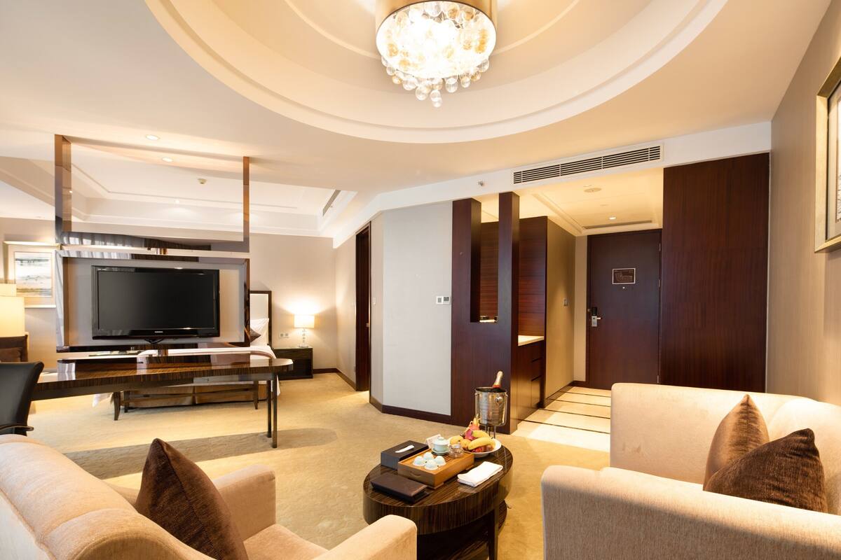 A modern hotel room with a flat-screen TV, a dining table, a sofa, and a chandelier.