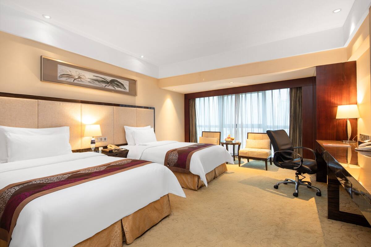 A hotel room with two beds, a desk, a chair, a lamp, and a window with curtains.