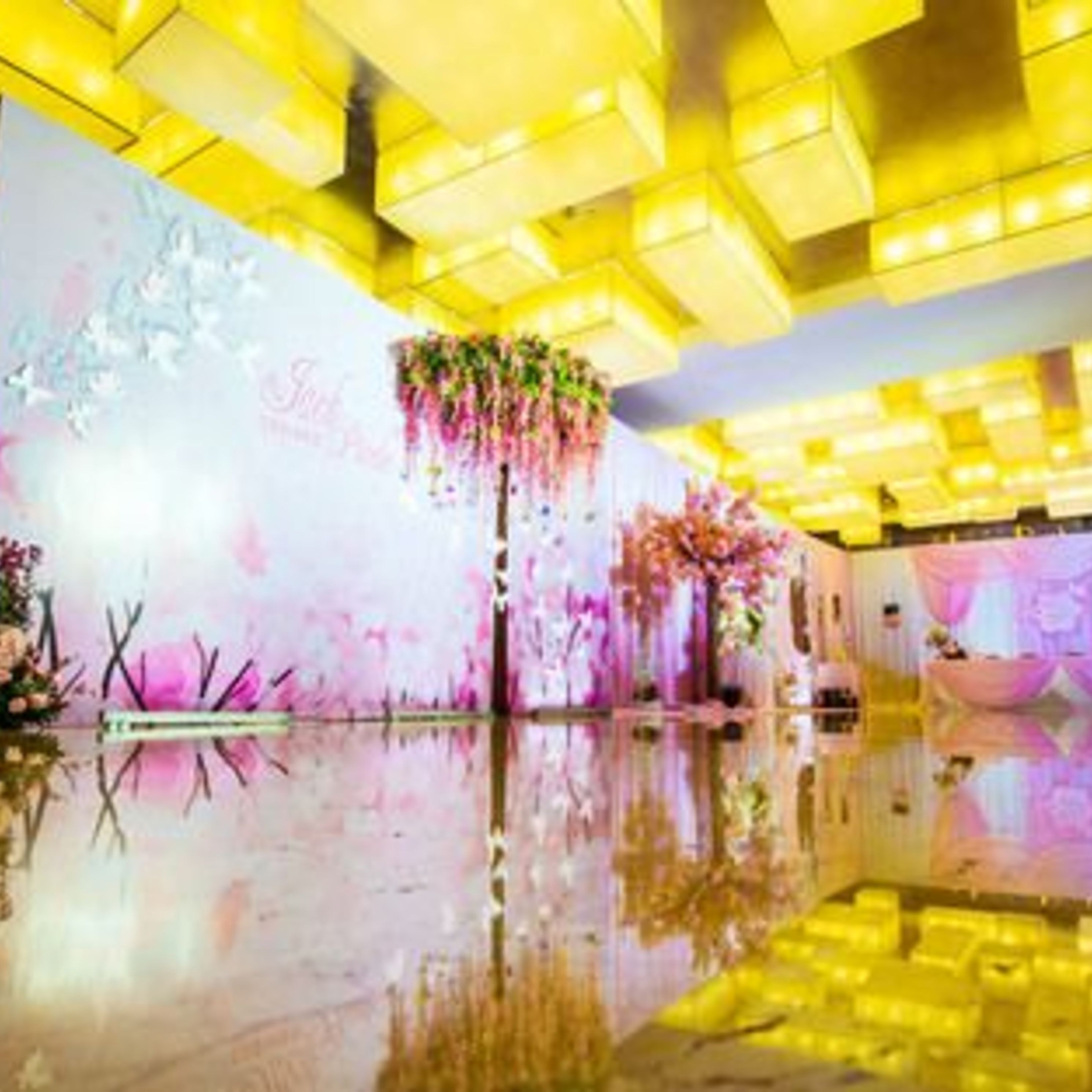A brightly lit indoor space with a reflective floor, a floral arrangement on the wall, and a ceiling with yellow lighting.