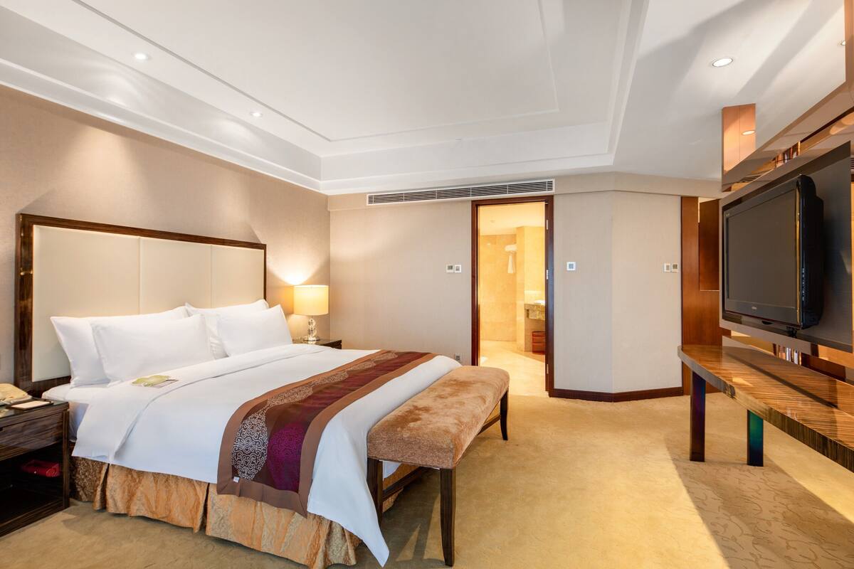 A hotel room with a large bed, bedside table, lamp, bench, TV, and a walk-in closet.