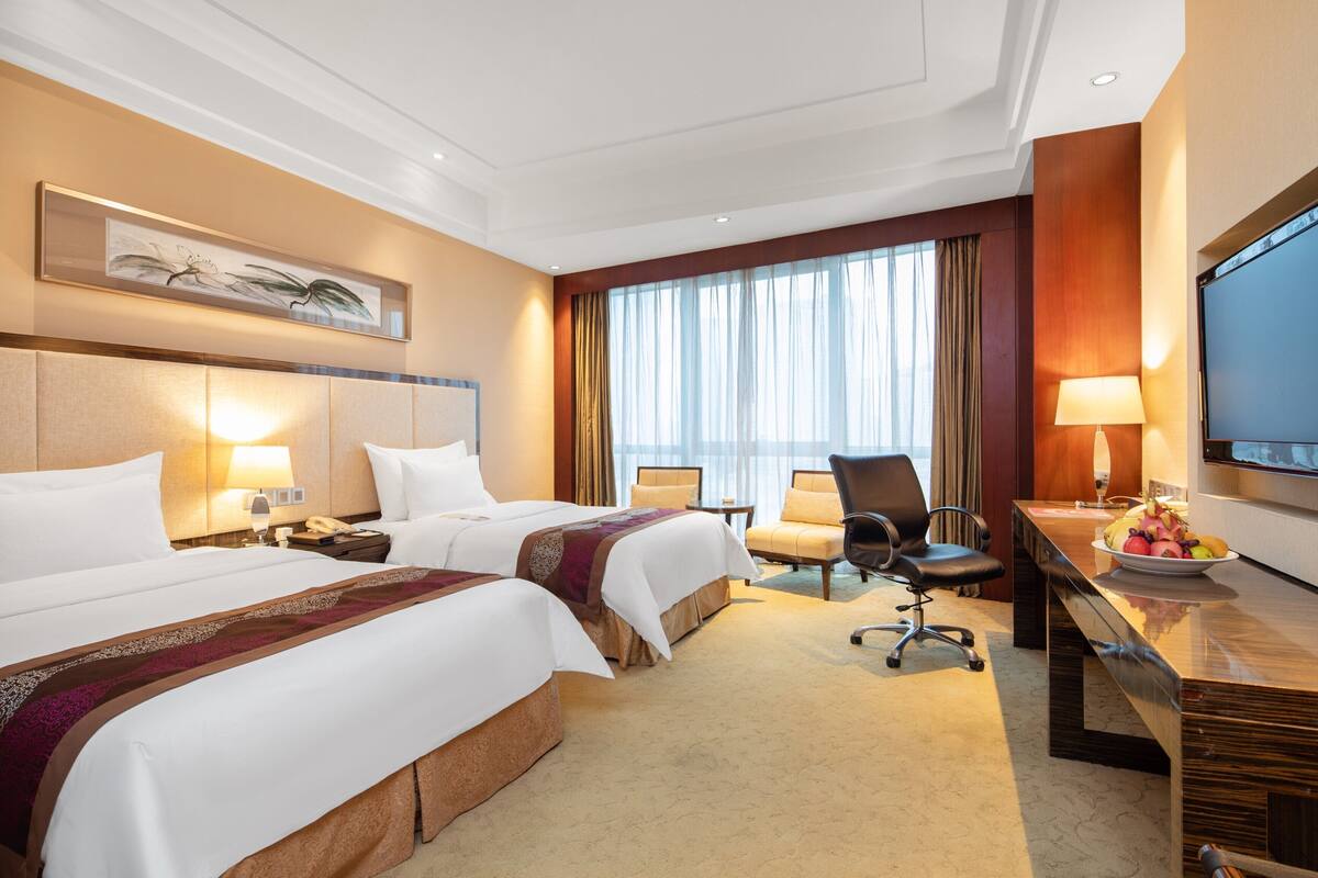 A hotel room with two beds, a desk with a television, a chair, and a large window with curtains.