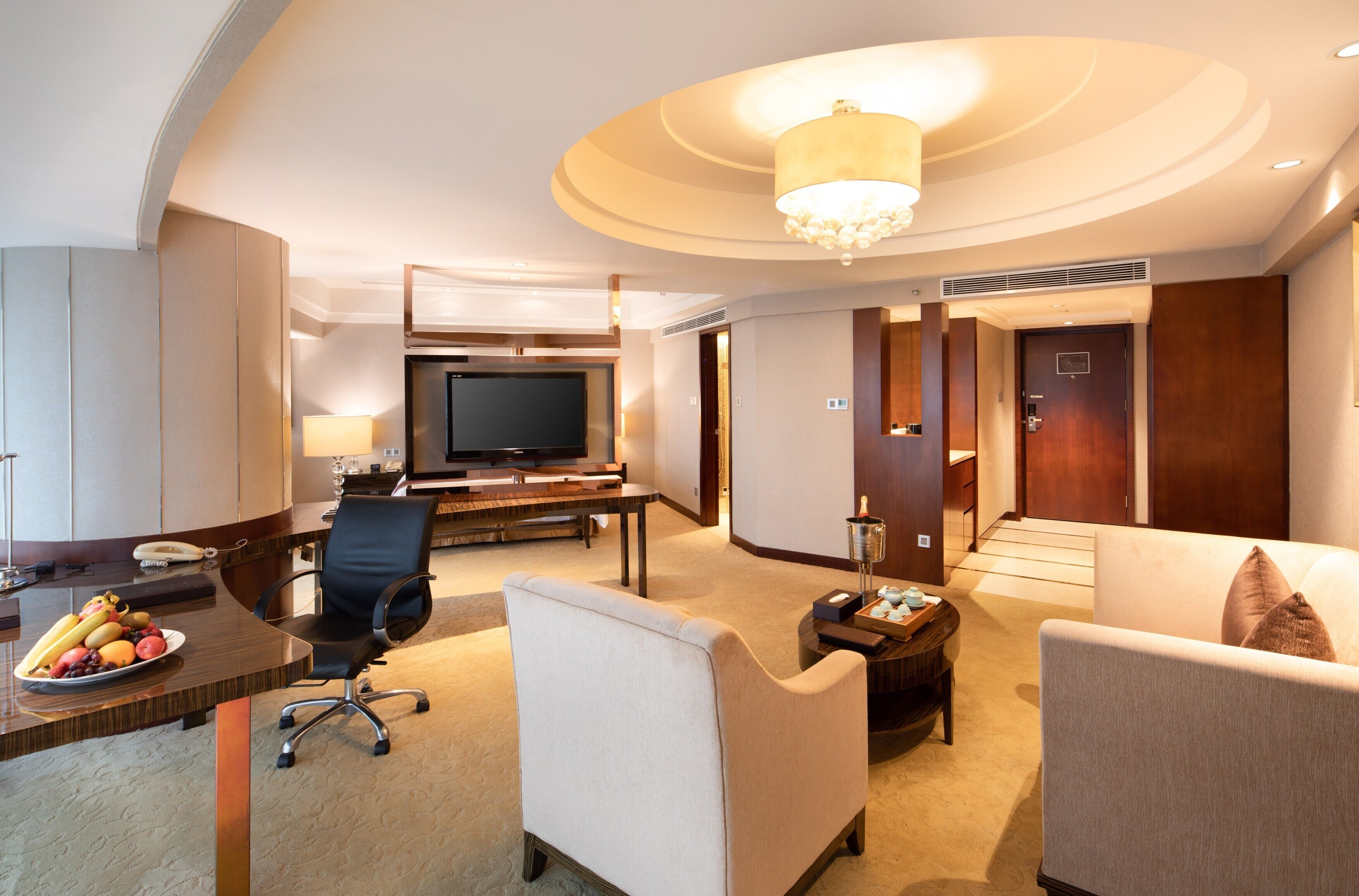 A hotel room with a flat-screen TV, a desk with a lamp, a dining table with fruit, and two sofas.