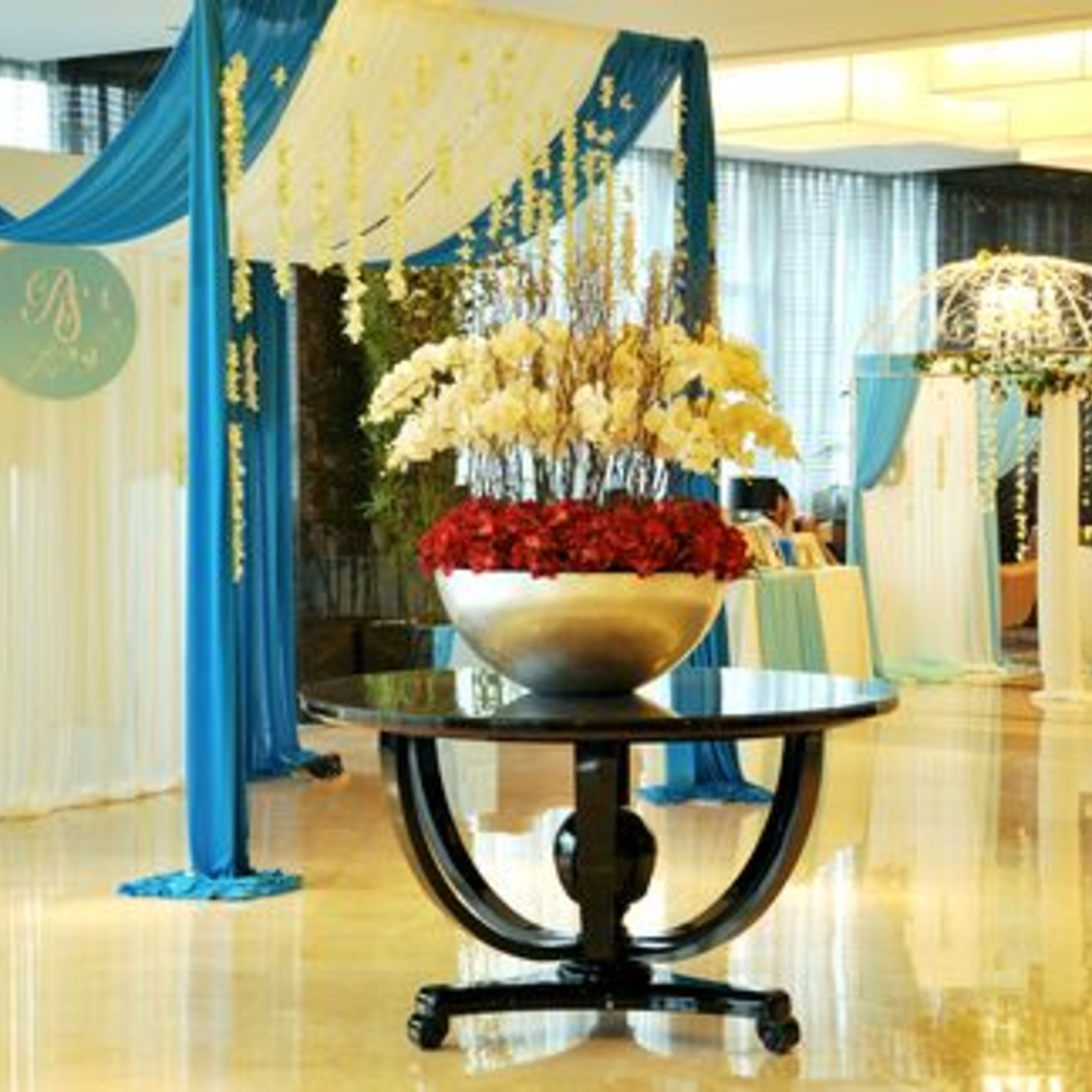 A table with a floral arrangement and a bowl of red flowers, set against a backdrop of blue drapes and a white wall.