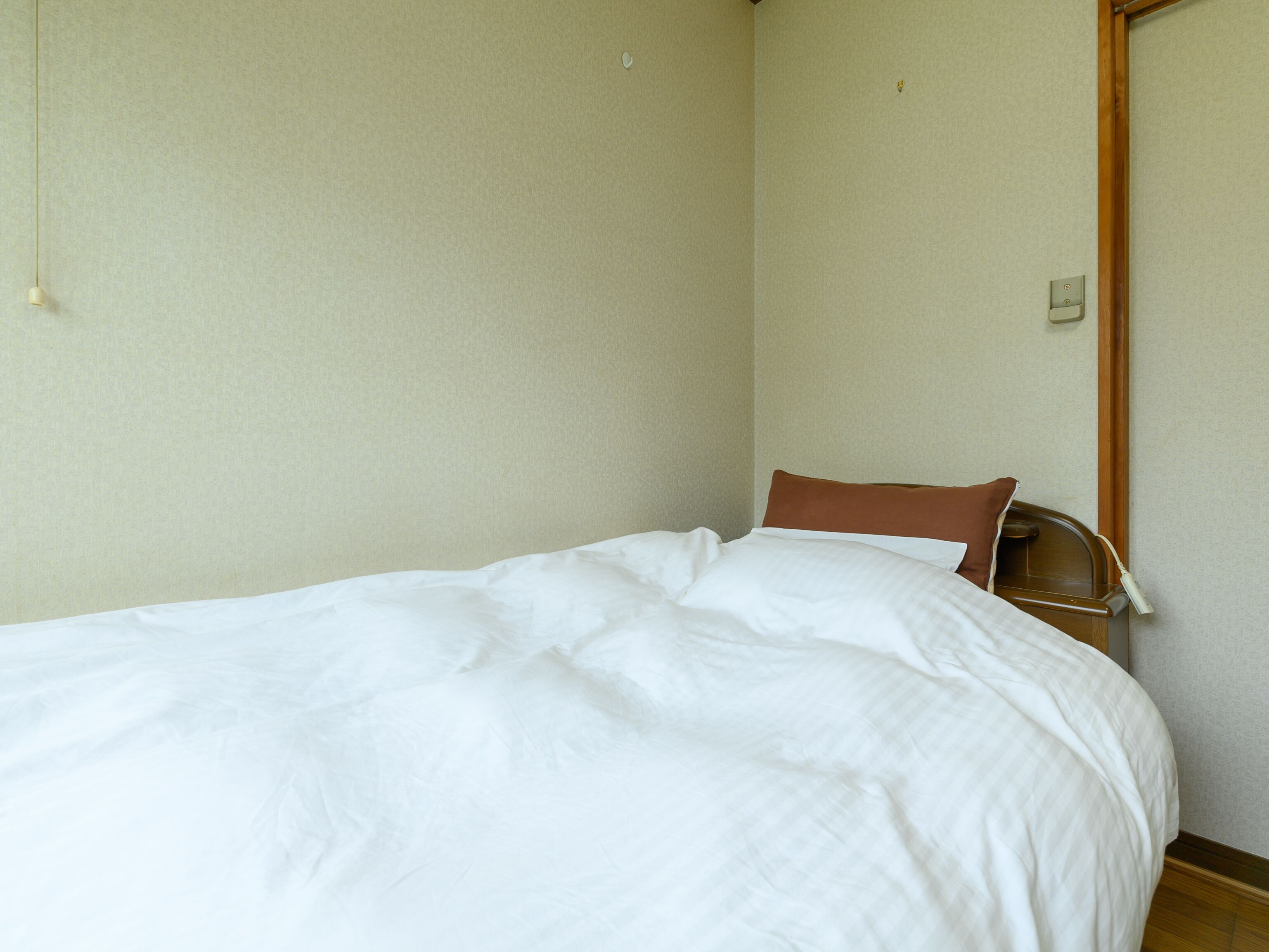 A single bed with white bedding in a room with plain walls and a wooden door frame.