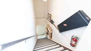 Una escalera estrecha con un extintor a la derecha, un cuadro en la pared y una lámpara colgante.