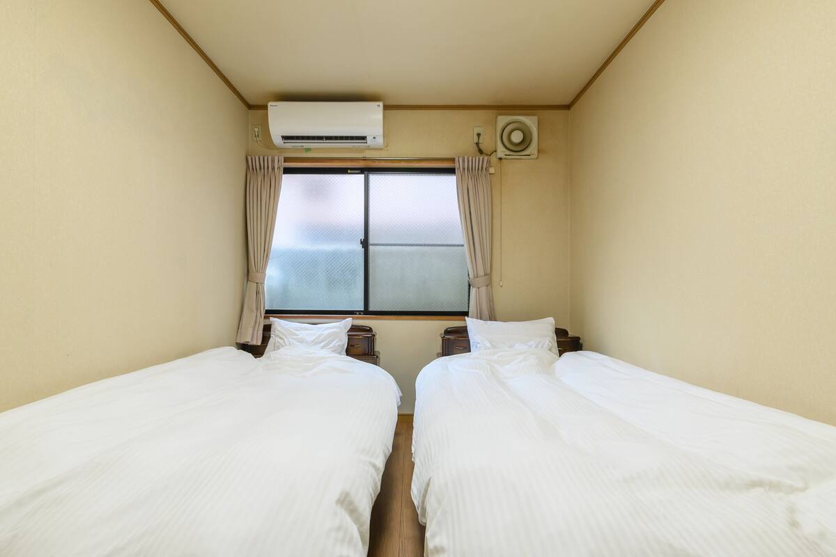 Habitación con dos camas, aire acondicionado, ventana con cortinas y ventilador de pared.