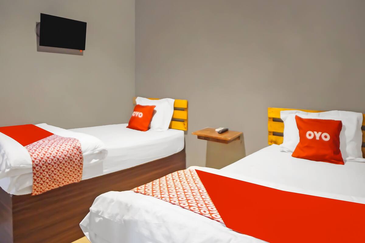 A hotel room with two beds, each with a red and white pillow labeled "OYO," a wooden nightstand, and a small table with a phone.