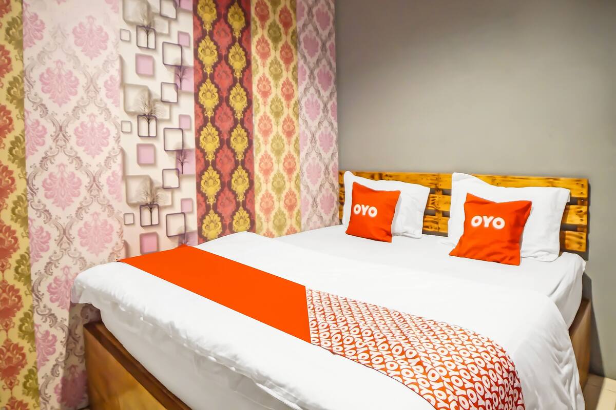 A neatly made bed with white and orange bedding, two pillows with the word "OYO" on them, and a patterned wall behind.