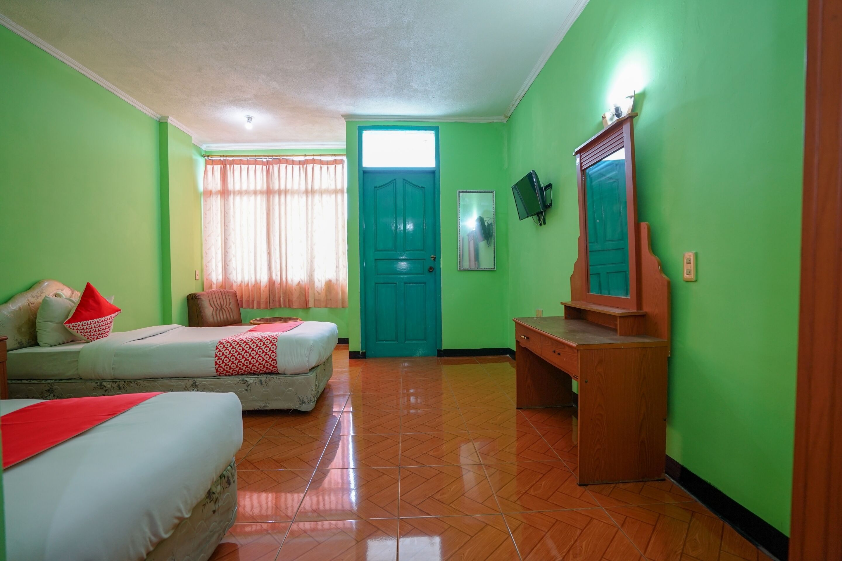 A room with two beds, a wooden desk, a mirror, and a green door.