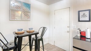 Een compacte kamer met een eettafel, twee stoelen en een klein kitchenette met een koelkast en koffiezetapparaat.