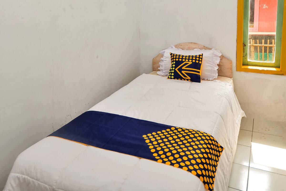 A single bed with a white and blue bedspread, a pillow with yellow dots and a zigzag pattern, and a window with a yellow frame.