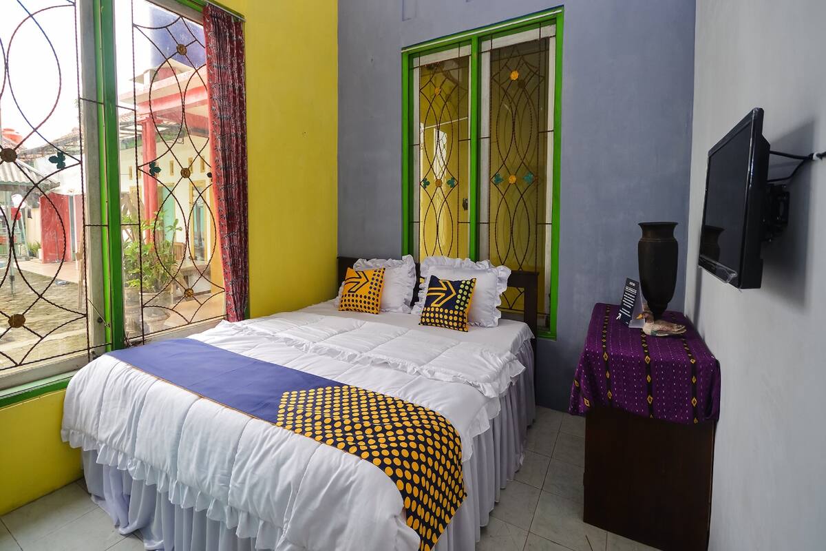 A bedroom with a bed, a window with green frames, a television, and a small table with a vase.