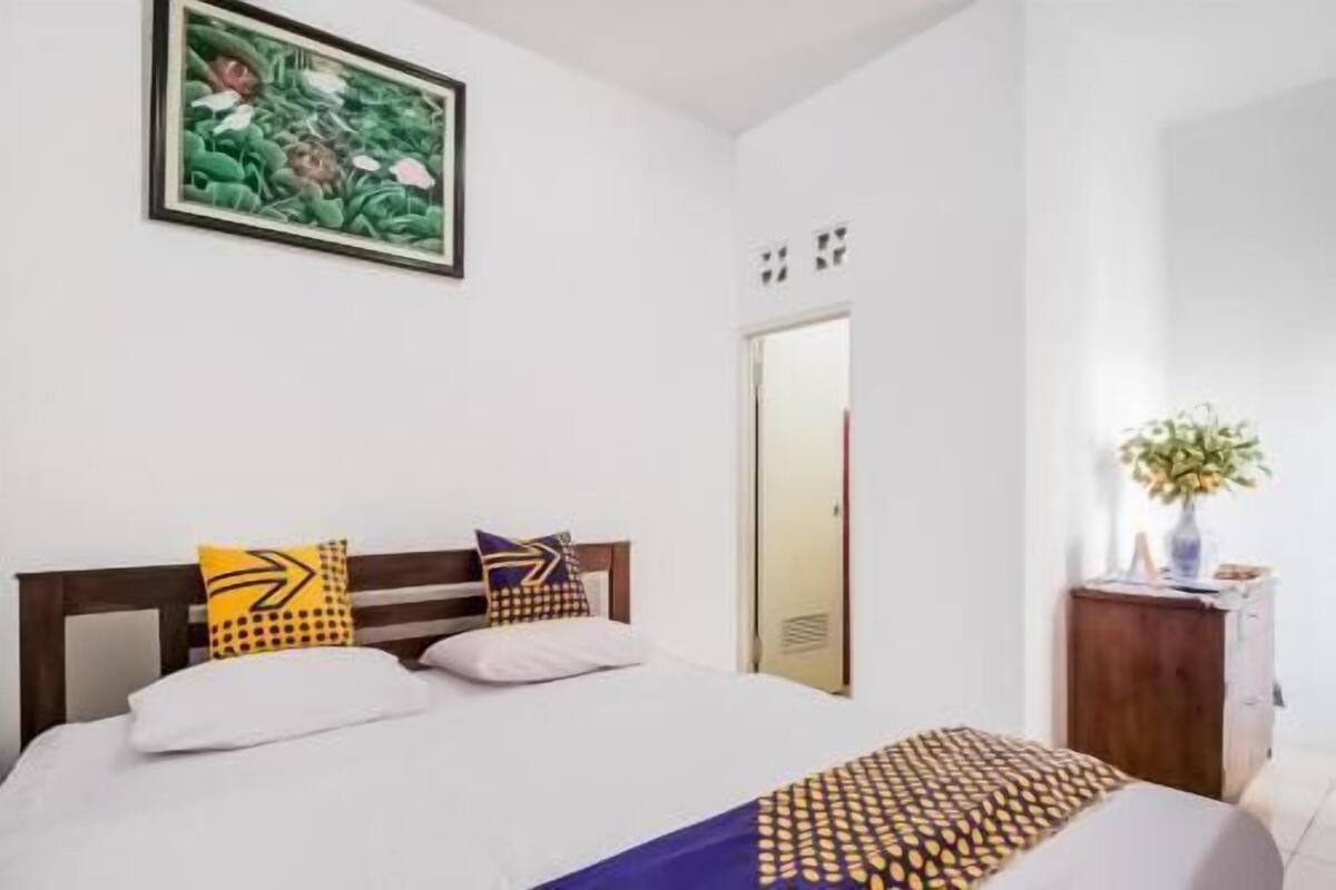 A bedroom with a bed, pillows, a painting, a mirror, and a vase with flowers.