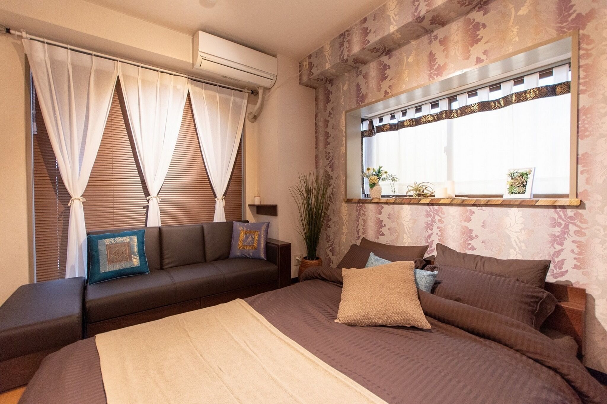 A bedroom with a bed, a sofa, a window with curtains, a potted plant, and a wall-mounted air conditioner.