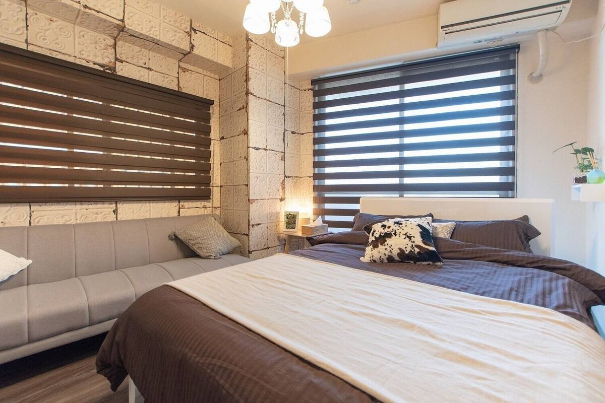 Un dormitorio con cama, sofá, mesita de noche, lámpara y ventana con persianas.
