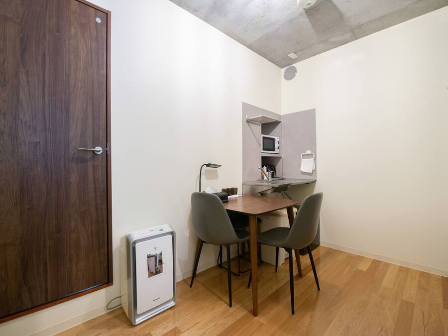 Une pièce de taille modeste avec une porte en bois, une petite kitchenette équipée d’un four à micro-ondes, une table avec deux chaises et un réfrigérateur compact.