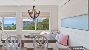 A dining area with a wooden table, white chairs, a bench with cushions, a chandelier, and large windows with a view of the ocean. - Gulf and State Park Views, Private Beach, HOT TUB (Santa Rosa Beach)