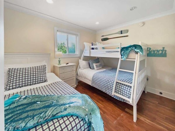 Une chambre avec deux lits, un lit superposé, une fenêtre, une table de chevet et une planche de surf fixée au mur.