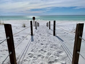Un sentier de sable qui longe la plage et mène à l’océan, avec des poteaux en bois et des cordes qui indiquent le chemin.