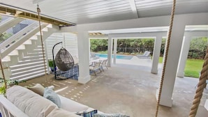 A covered outdoor seating area with a swing chair, a cushioned bench, and a view of a pool and garden. - 30A Beachfront and State Park Views, Gulf Trace (Santa Rosa Beach)