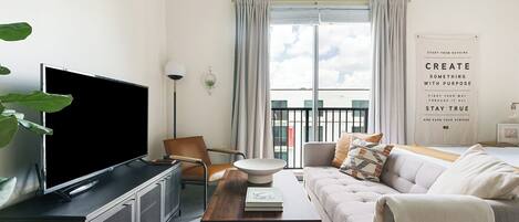 A modern living room with a flat-screen TV, a wooden coffee table, a sofa, and a large window with curtains.