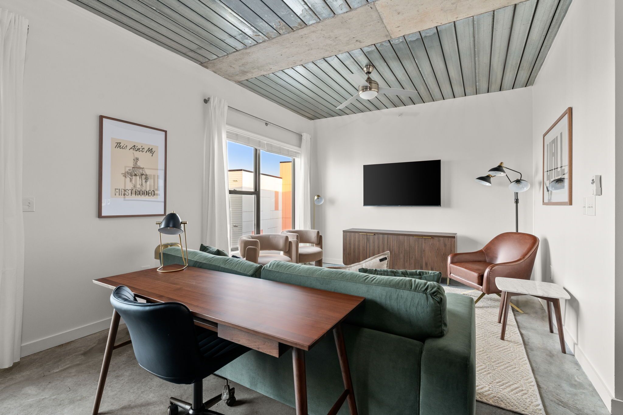 A modern living room with a wooden dining table, green sofa, and a brown leather chair. A flat-screen TV is mounted on the wall, and a ceiling fan is visible.