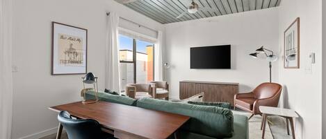 Una sala de estar moderna con una mesa de comedor de madera, un sofá verde y un sillón de cuero marrón. Un televisor de pantalla plana está montado en la pared, y se observa un ventilador de techo.