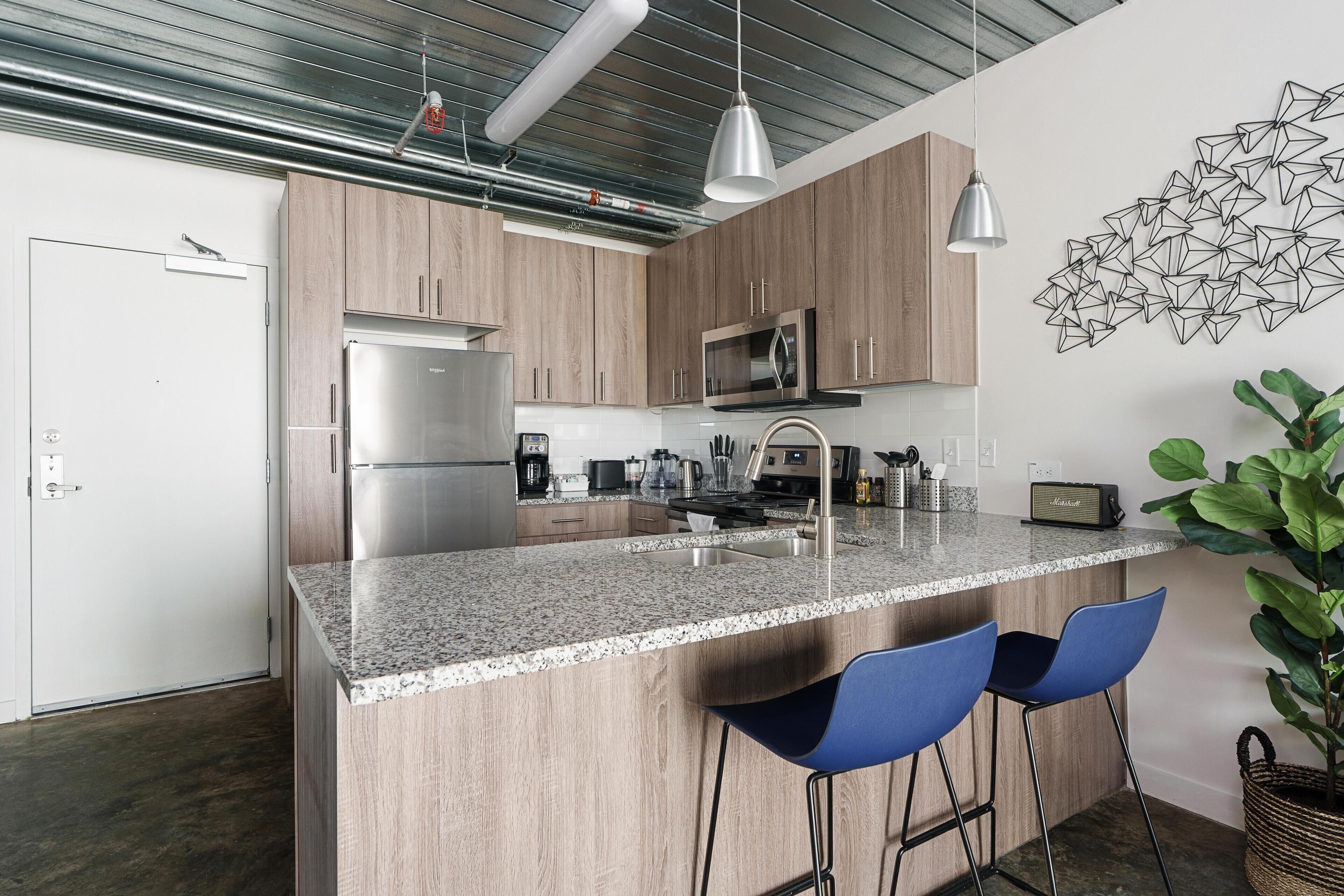 A modern kitchen with a granite countertop, stainless steel appliances, and blue bar stools.