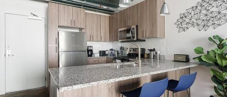 A modern kitchen with a granite countertop, stainless steel appliances, and blue bar stools.