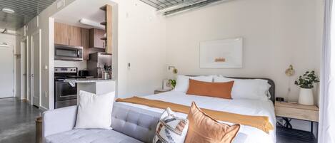 A modern hotel room with a sofa, a bed with a wooden headboard, a nightstand, a wall-mounted TV, and a kitchen area with appliances.