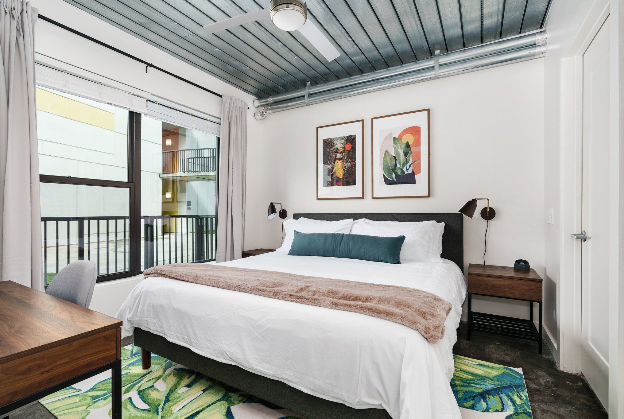 A modern bedroom with a large bed, a desk, and a view of the outside balcony.