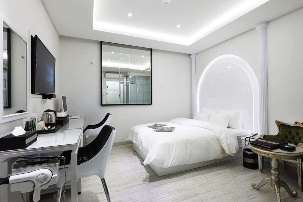 Habitación de hotel moderna con cama, escritorio con computadora, silla, tocador con espejo y televisor.