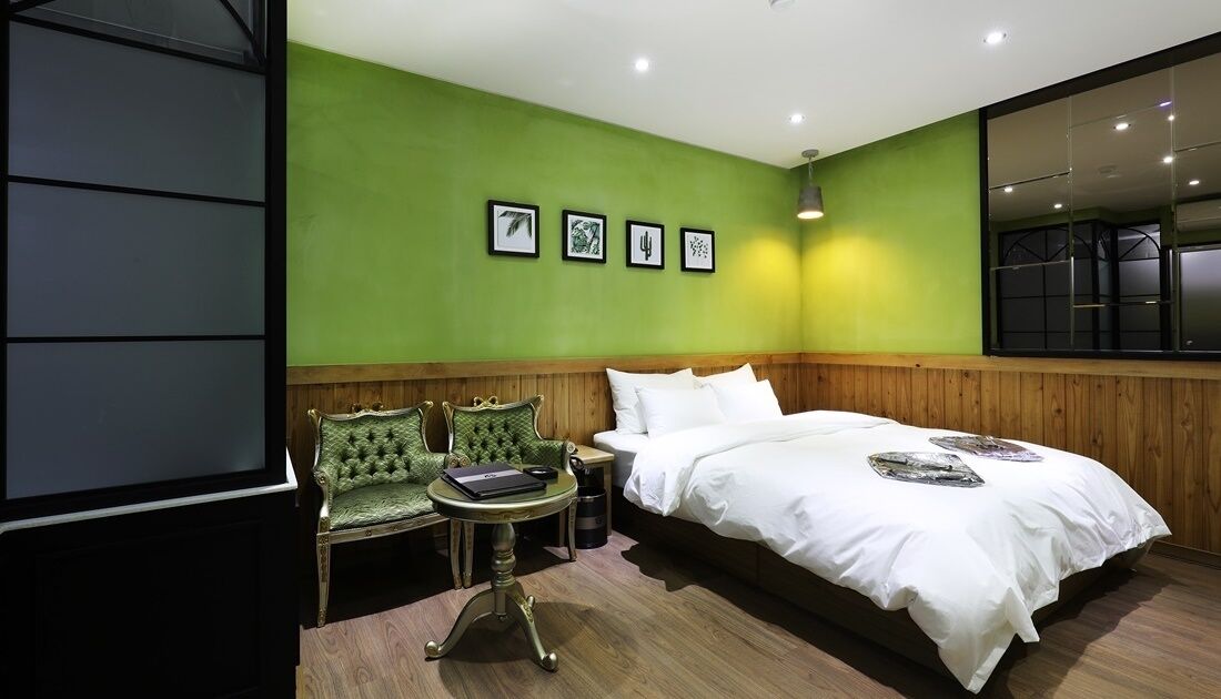 A hotel room with a green wall, a large bed, a wooden headboard, a green armchair, a small table, and a mirror.