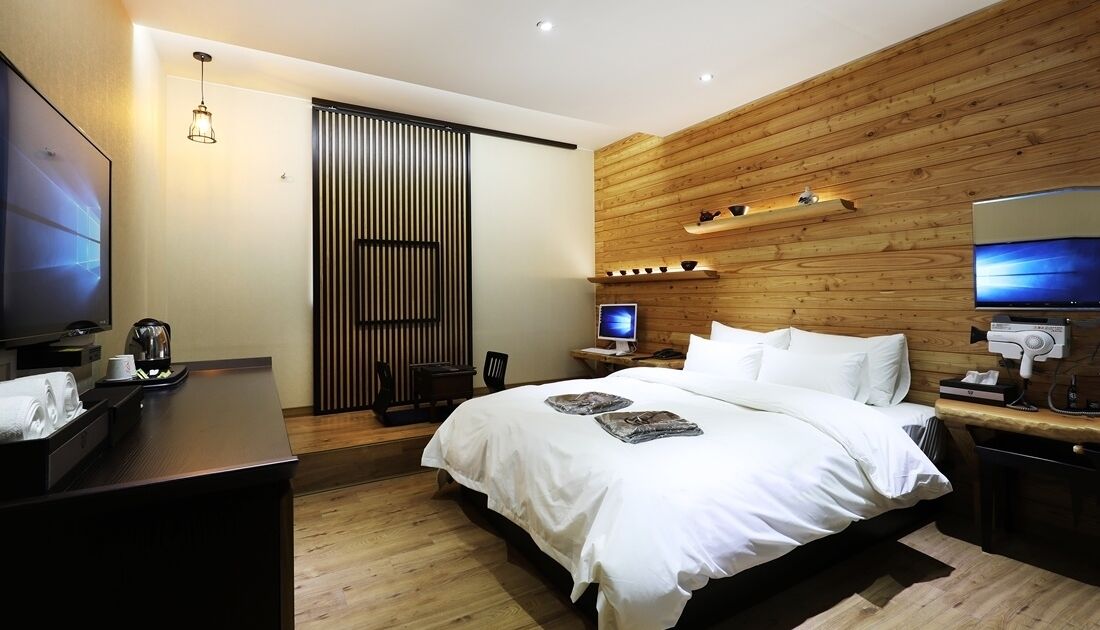 A modern hotel room with a large bed, a desk with a computer, a TV, and a wooden wall panel.