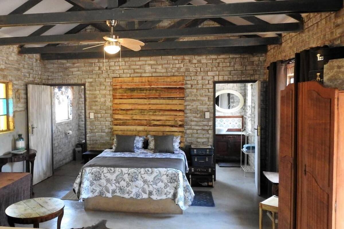 A rustic room with exposed brick walls, a wooden ceiling, a bed with floral bedding, a small table, and a window with stained glass.