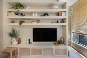 A living room with a flat-screen TV mounted on the wall, surrounded by shelves with books, decorative items, and plants.