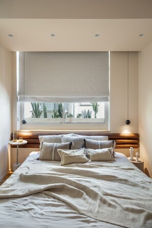 A bedroom with a large bed, a window with a view of plants, a bedside table with a lamp, and a ceiling light.