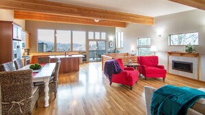 A spacious living room with a wooden ceiling, a dining area with wicker chairs, a red armchair, and a fireplace. - Blackberry Bramble - Beckoning Views, Peaceful Elegance, Walk to Downtown (Hood River)