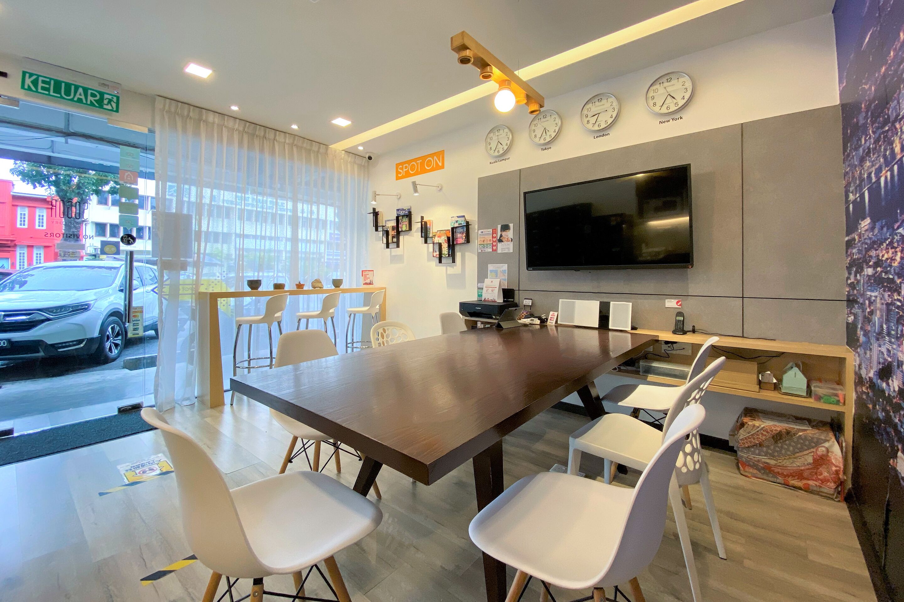 A modern hotel room with a large dining table, white chairs, a flat-screen TV, clocks on the wall, and a view of the street outside.