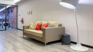 Eine moderne Hotellobby mit einem beigen Sofa, roten und beigen Kissen, einer weißen Stehlampe und einem sichtbaren Feuerlöscher.