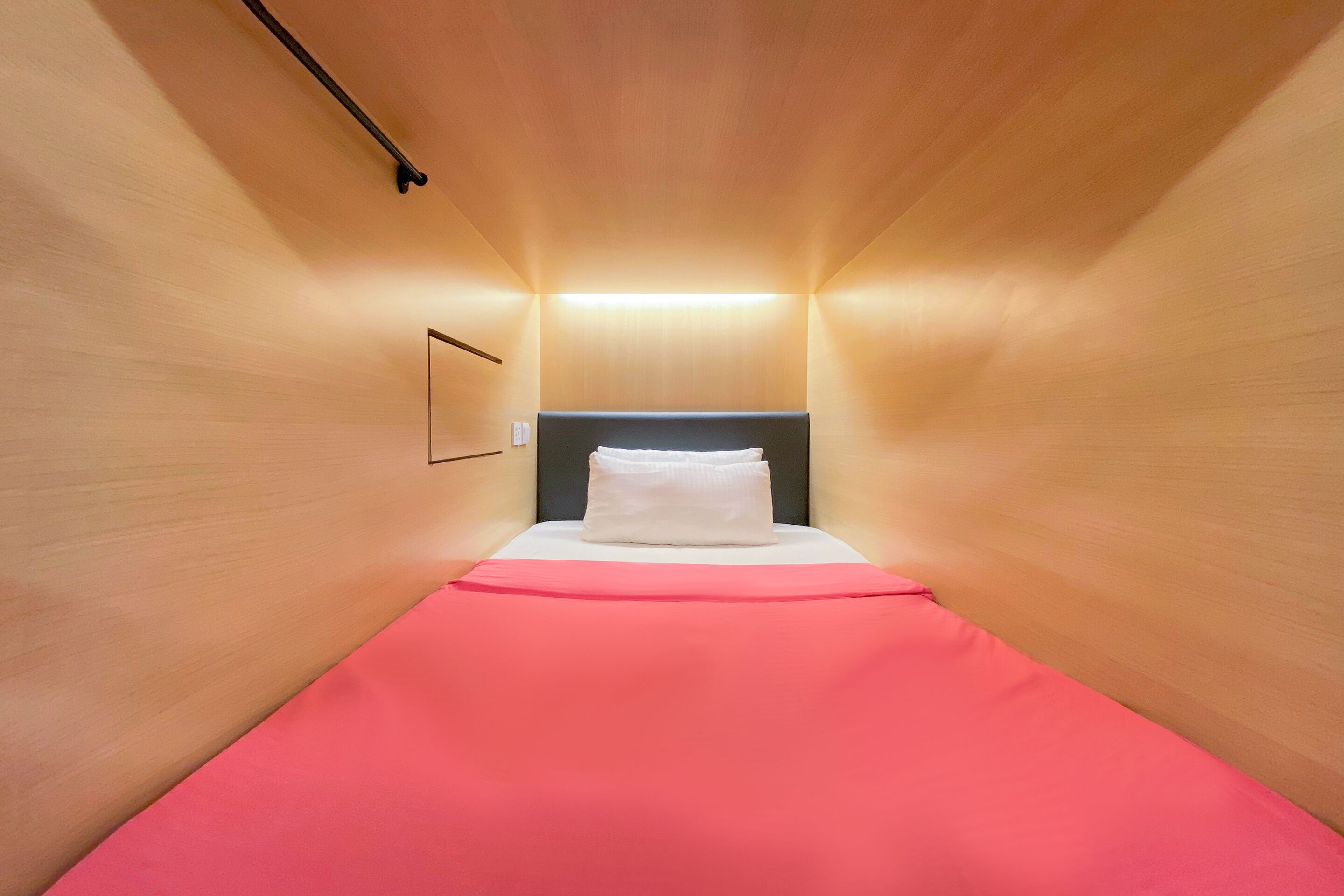 A narrow, wooden-paneled room with a bed centered, a headboard, and a wall-mounted light fixture.