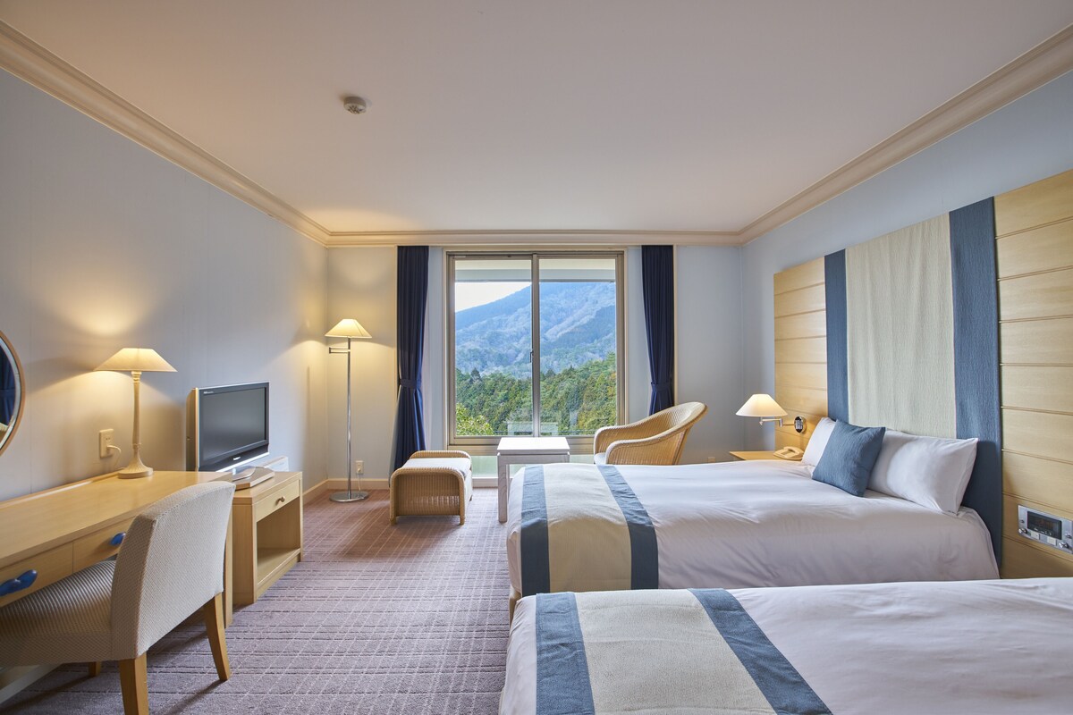 Hotelkamer met twee bedden, een bureau met een televisie, een stoel, een kleine tafel en uitzicht op bergen door het raam.