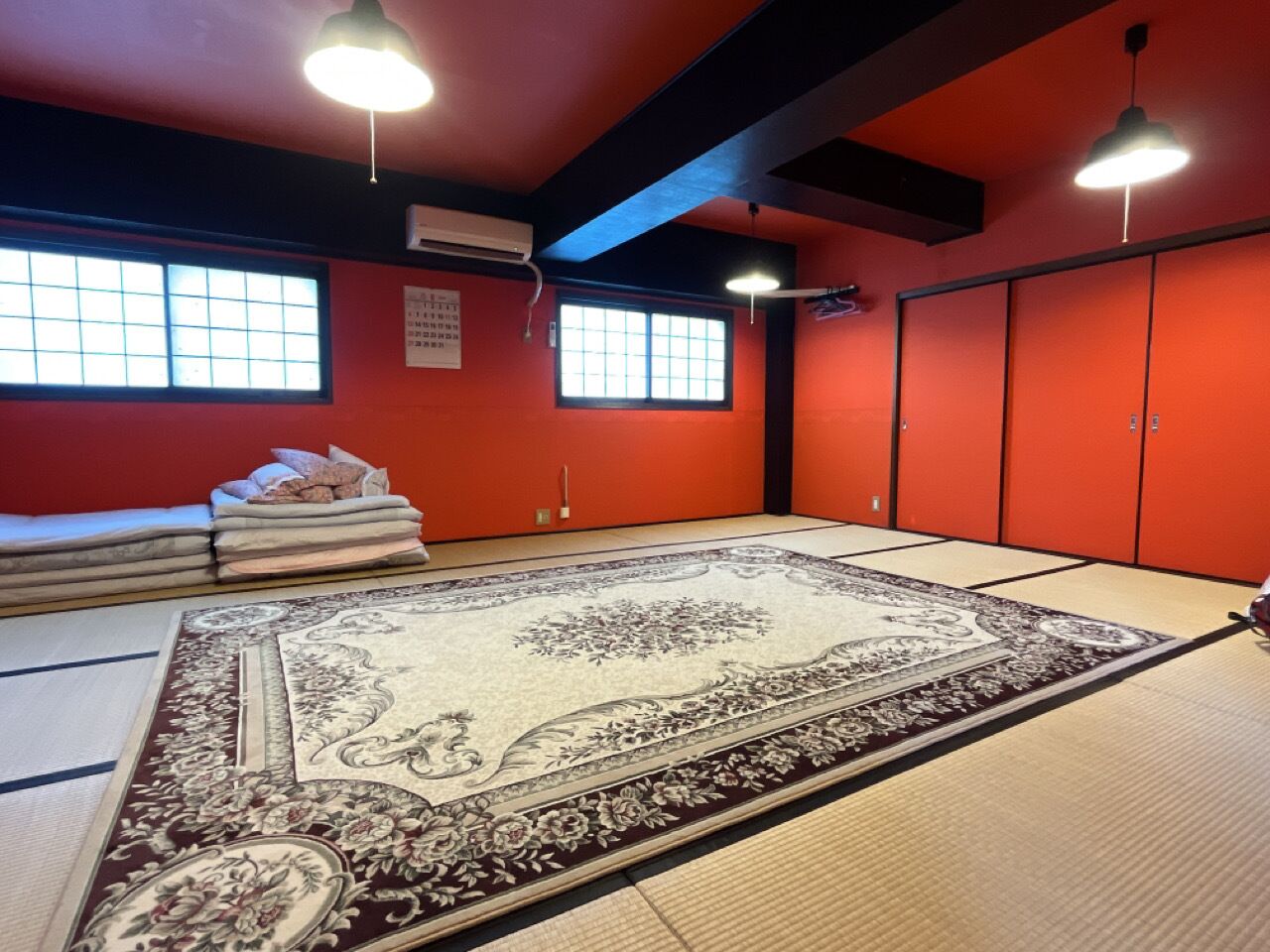 A tatami mat room with a large decorative rug, a window, and a stack of folded mats.